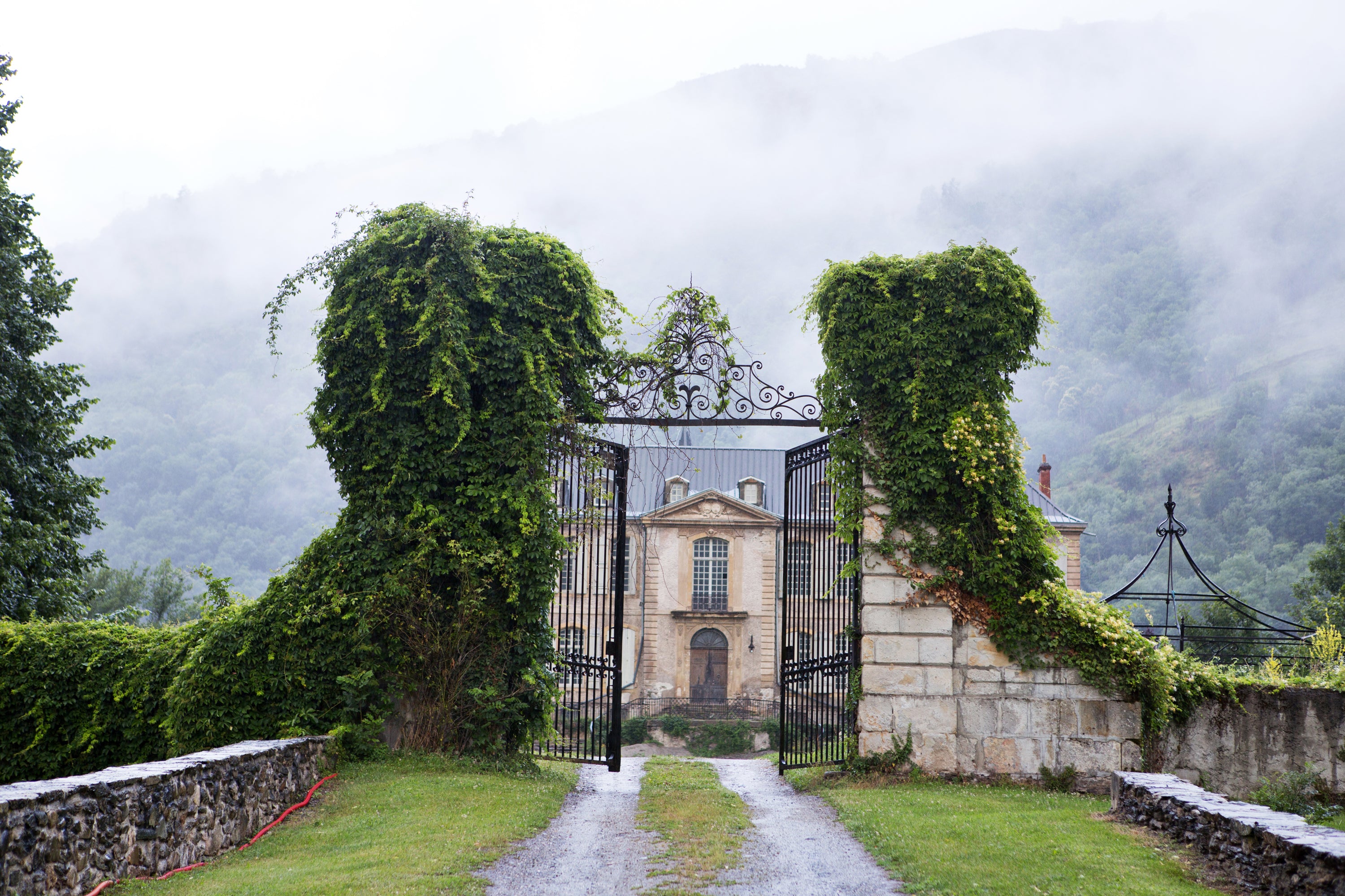 Majestic French chateau with a grand gate surrounded by lush greenery and misty mountains — editorial property photography by Carla Coulson