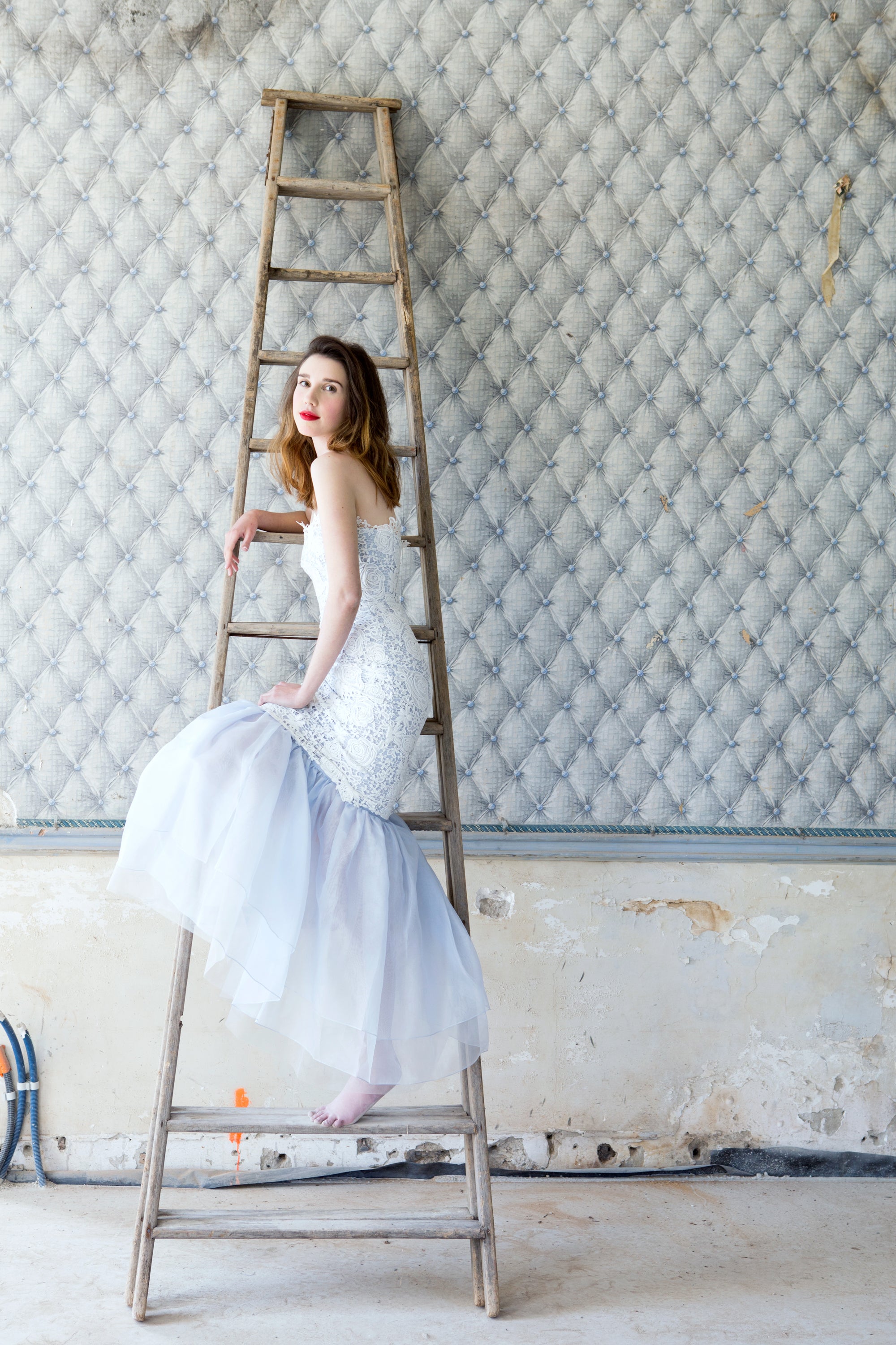 Woman in a white dress sitting on a ladder against a textured wall in a French chataeu — editorial photography by Carla Coulson